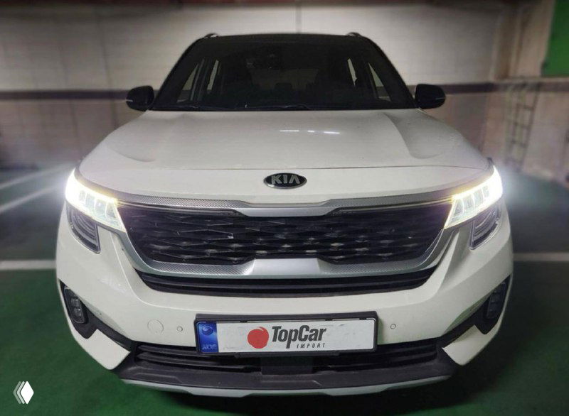 Фронтальный снимок белого KIA Seltos с включёнными дневными ходовыми огнями, видна решётка радиатора и эмблема Kia на капоте в гаражных условиях.