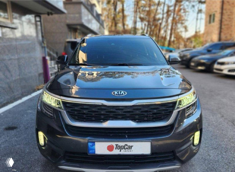 Чёрный KIA Seltos на улице, отражение зданий на капоте, видна передняя часть кузова, противотуманные фары и фирменная решётка радиатора Kia.