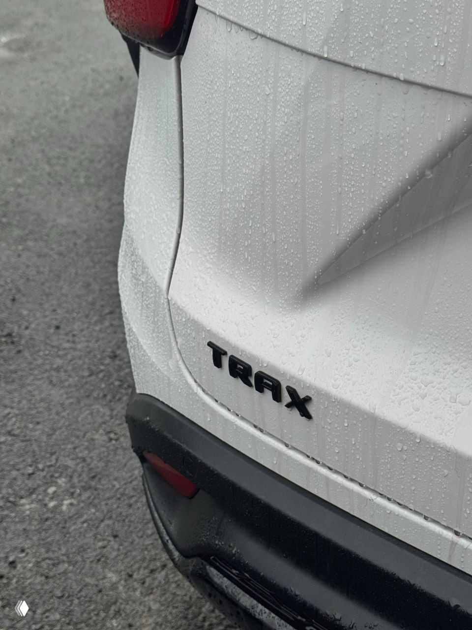 Крупный план задней части автомобиля с шильдиком TRAX, белый лак покрыт каплями воды после дождя, видна часть бампера