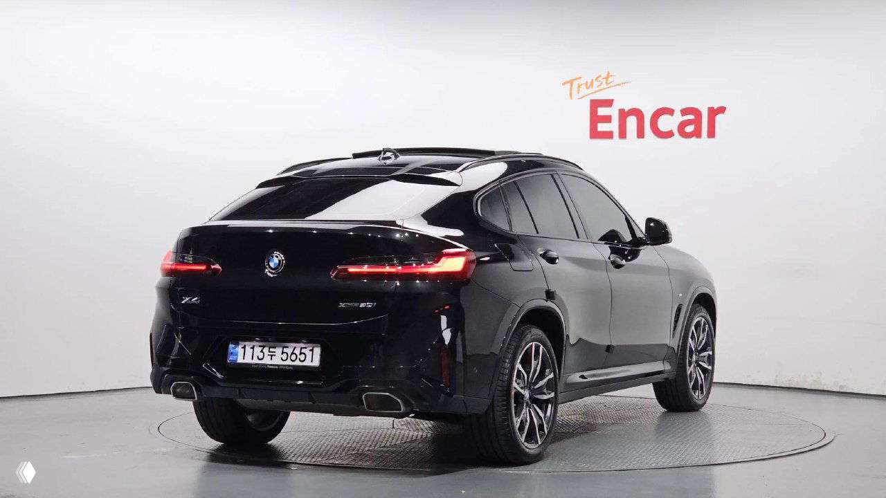 Задний ракурс чёрного BMW X4 xDrive 20i M‑Sport в шоуруме, видно задние фонари, номер в корейском формате и выхлопную систему на парковочном диске.