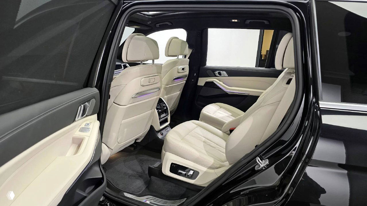 Задние пассажирские кресла BMW X7 с бежевой кожей, пространство для ног и элементы подсветки интерьера на фоне открытой задней двери.