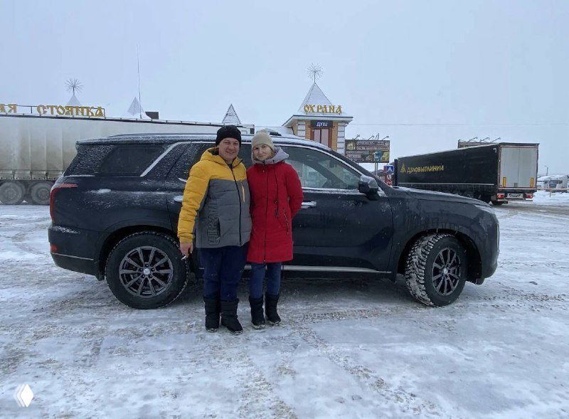 Два человека стоят рядом с чёрным Hyundai Palisade на снежной стоянке — фото клиента и автомобиля после доставки в Россию.