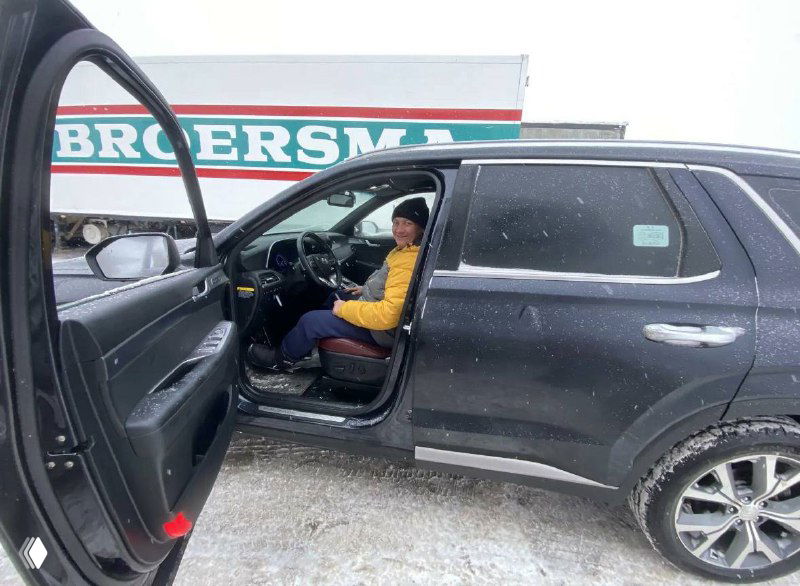 Клиент сидит в водительском кресле Hyundai Palisade с открытой дверью, видно салон и часть кузова на фоне парковки и грузовиков.