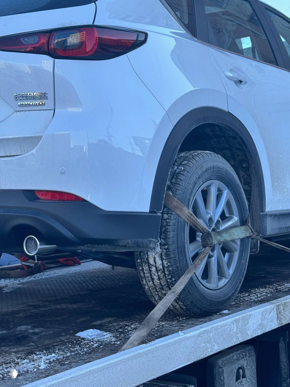 Крупный план заднего колеса Mazda CX-5: автомобиль зафиксирован стропами на платформе эвакуатора, видны следы снега и сцепления ремней.