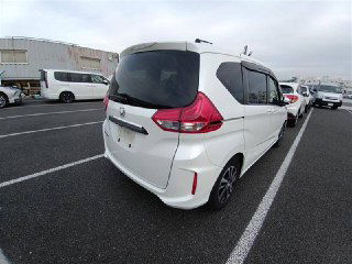 Вид сзади и боковой ракурсы белого Honda Freed 2018 на парковке аукциона: видно задние фонари, состояние лакокрасочного покрытия и соседние автомобили.