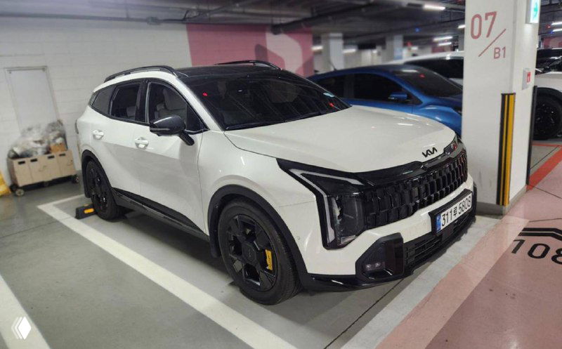 Угол спереди и сбоку белого KIA Sportage 2025 в паркинге, видны чёрные акценты на колёсах, защита порогов и стилистика бампера.