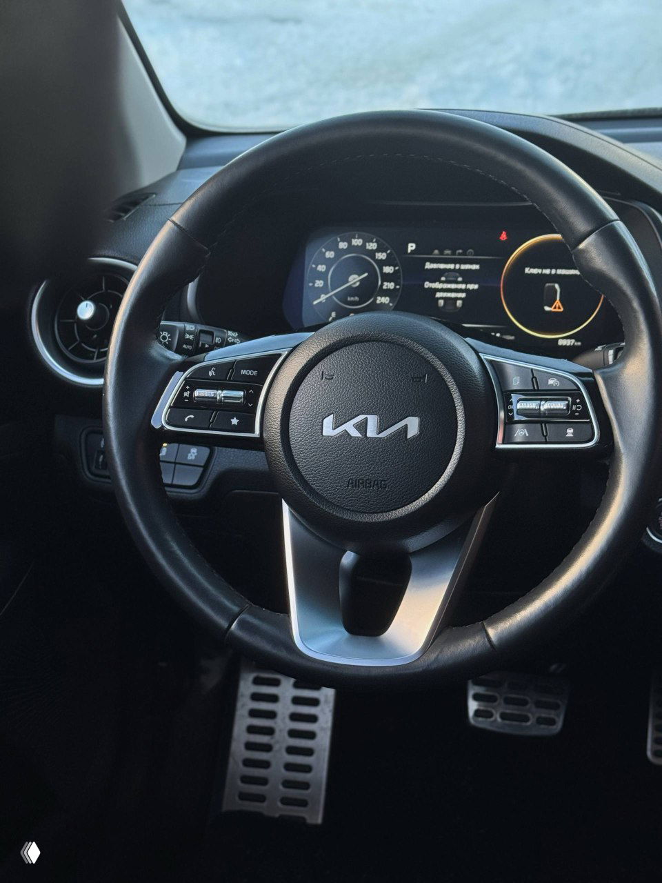 Рулевое колесо KIA с логотипом, крупный план на кнопки управления аудио и круиз-контролем, цифровая приборная панель на фоне.
