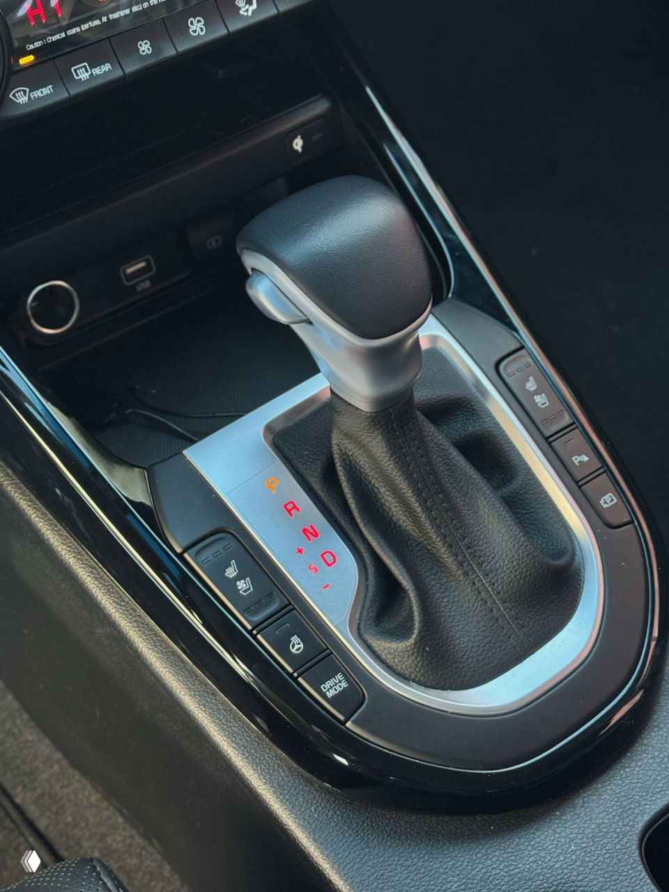 Рычаг вариатора KIA K3 внизу центральной консоли, рядом кнопки подогрева сидений и режимов движения, чистая обшивка вокруг.