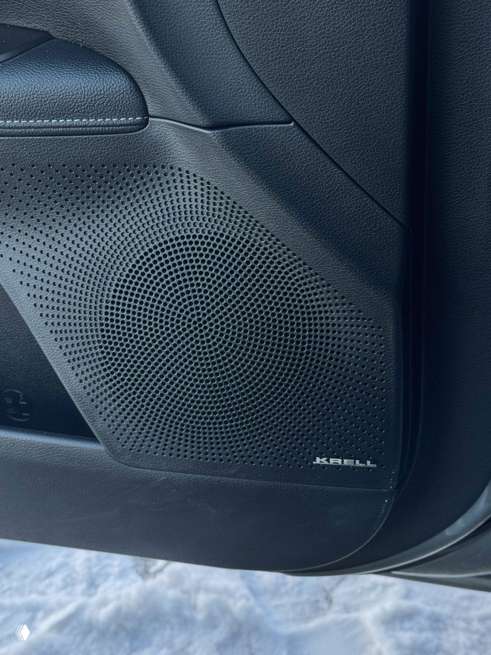 Крупный план звуковой решётки динамика с логотипом KRELL в нижней части дверной карты, часть порога и снег под машиной.