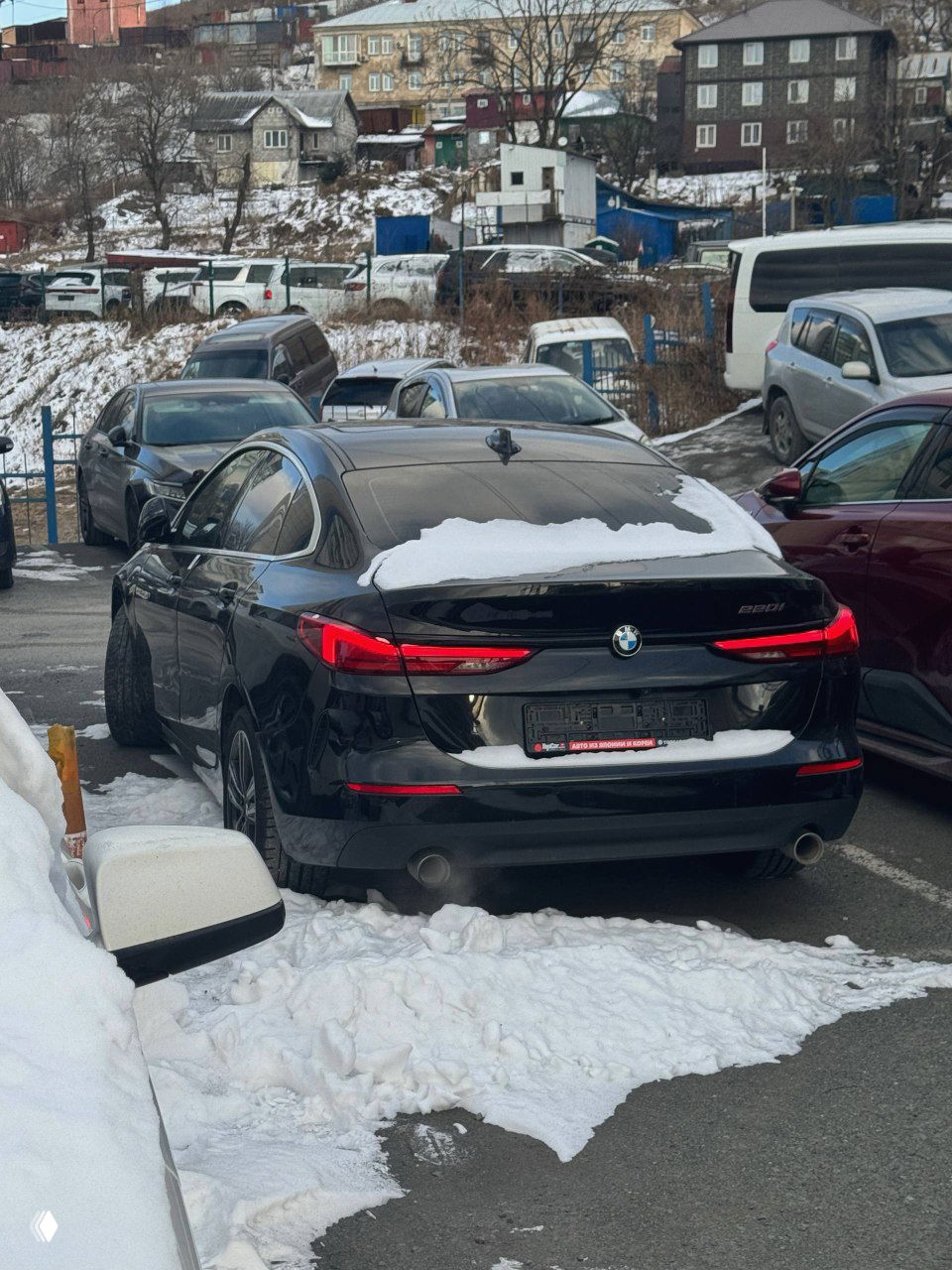 Задний вид чёрного BMW 220i 2021 года на стоянке, заметен снег на крышке багажника и номерной держатель, городской фон и припаркованные авто.