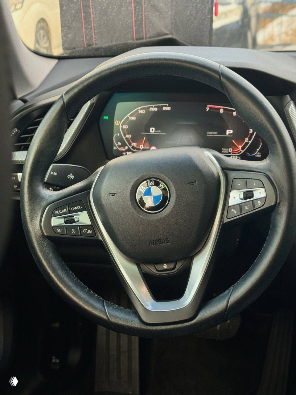 Крупный план рулевого колеса BMW с эмблемой, кнопками управления и фрагментом цифровой приборной панели, состояние и отделка салона видны ясно.