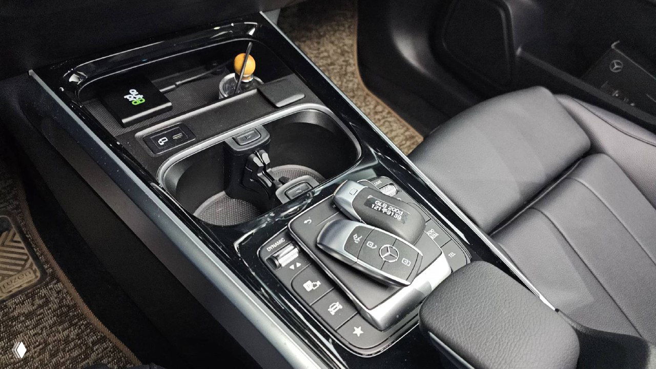 Центральный тоннель Mercedes GLB200d: кнопки управления, подстаканники и ключ-брелок Mercedes на декоративной панели, чёрная лакированная отделка.