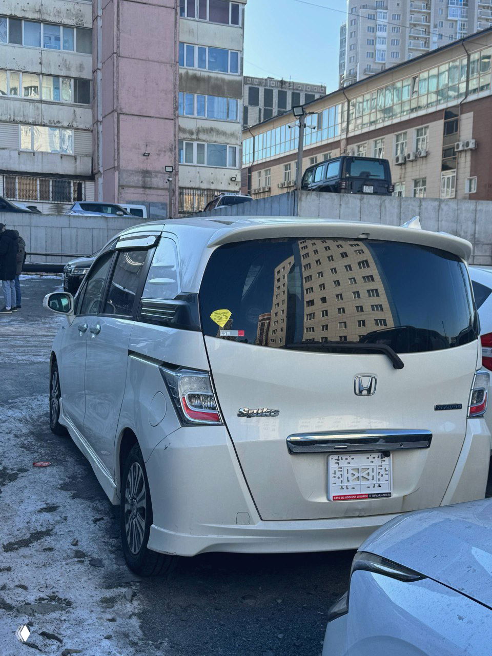 Задняя часть белого Honda Freed с правым рулём, вид на багажную дверь и задние фонари, автомобиль припаркован на заснеженной площадке.