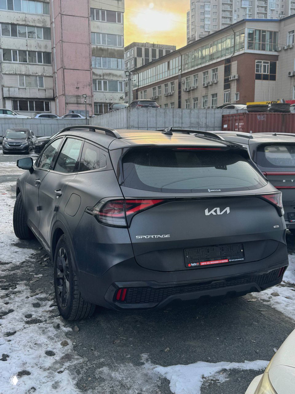 KIA Sportage 2023 вид сзади: багажник и задняя оптика, номера демонтированы, автомобиль на городской парковке рядом с другими машинами и сугробами.