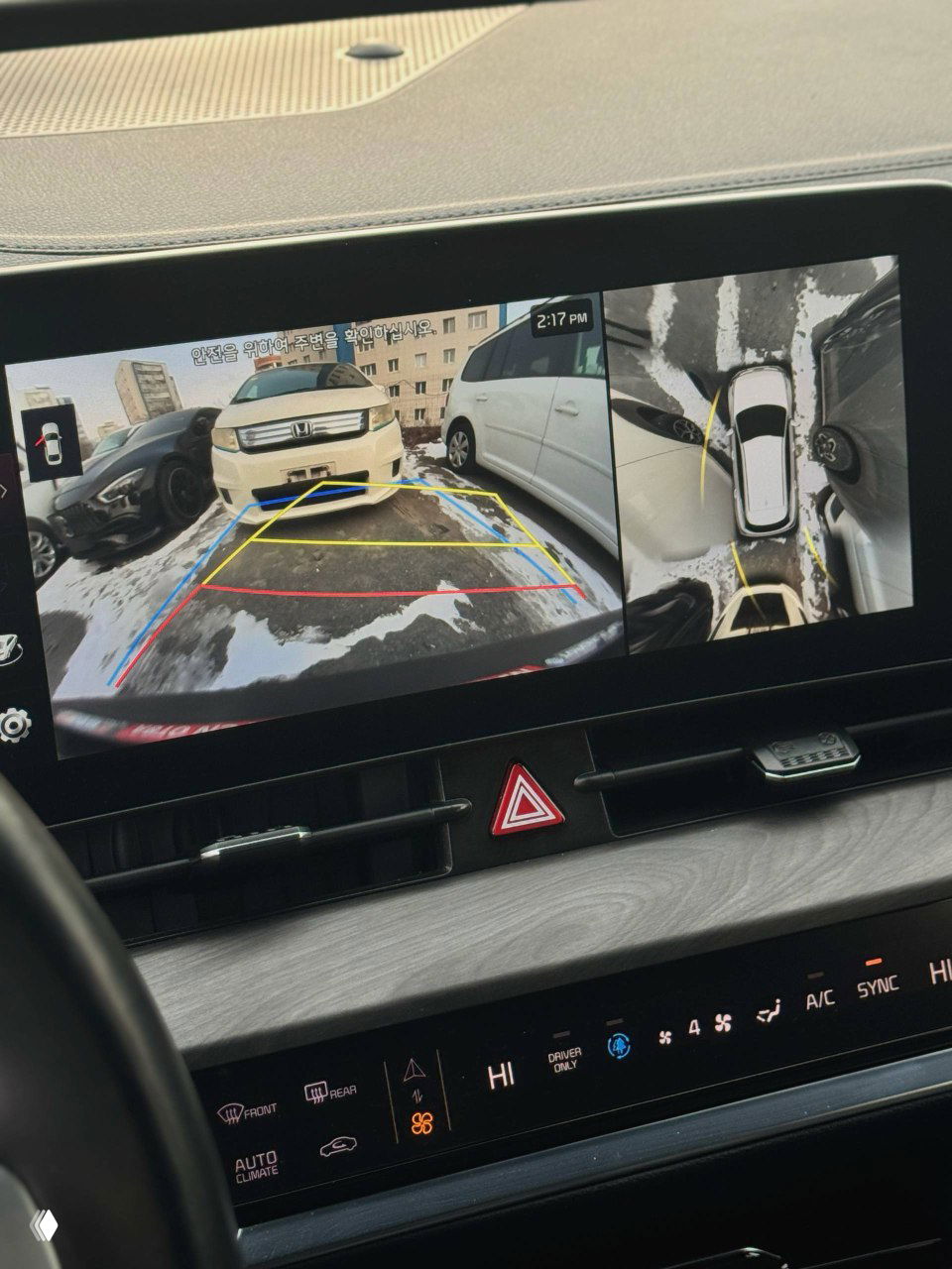 Экран камеры заднего вида с разметкой парковки и обзором сверху, видно другие припаркованные автомобили и снежную лужу, изображение системы помощи при парковке.