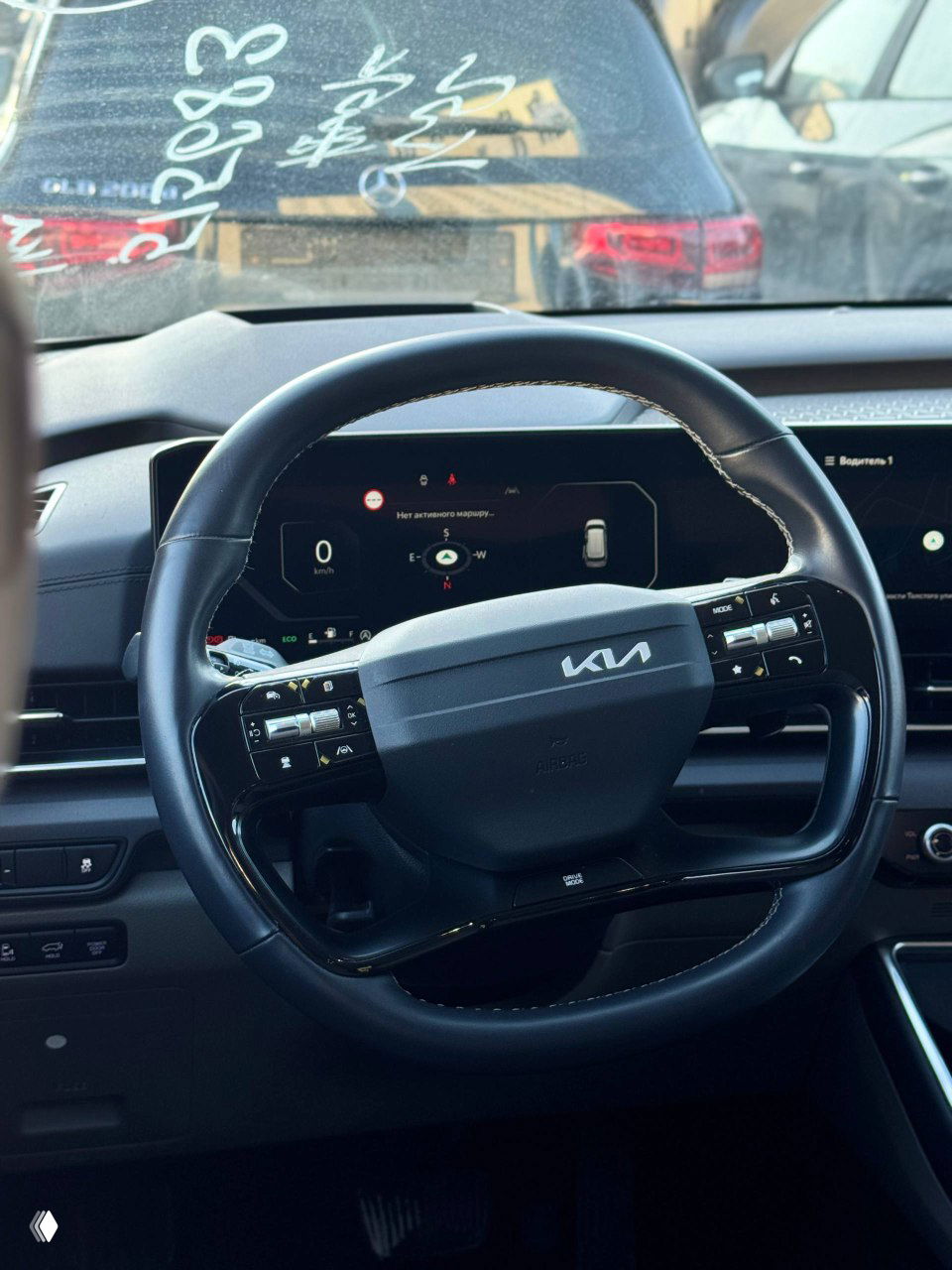 Вид рулевого колеса KIA с логотипом KIA и цифровой приборной панелью, кадр сделан из салона через лобовое стекло.