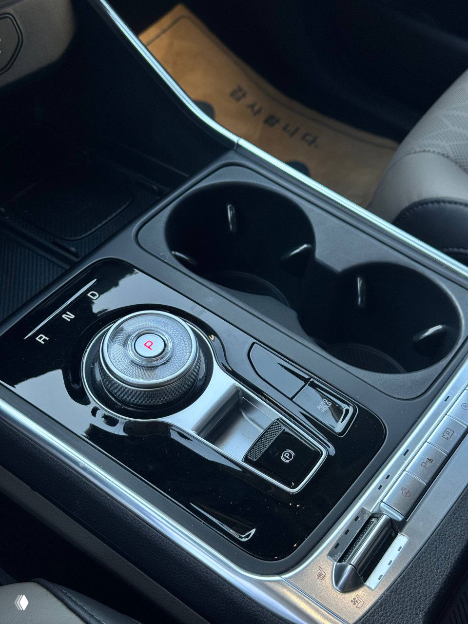 Фрагмент центральной консоли KIA Carnival: селектор-руль с вращающимся рычагом передач, подстаканники и кнопки управления на глянцевой панели.