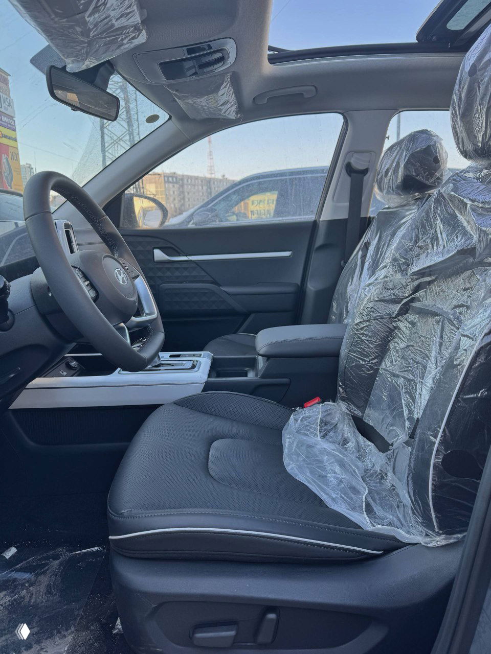 Переднее пассажирское сиденье Hyundai Mufasa в защитной плёнке, видно кресло, подлокотник и панорамный люк над салоном на фоне городской застройки.