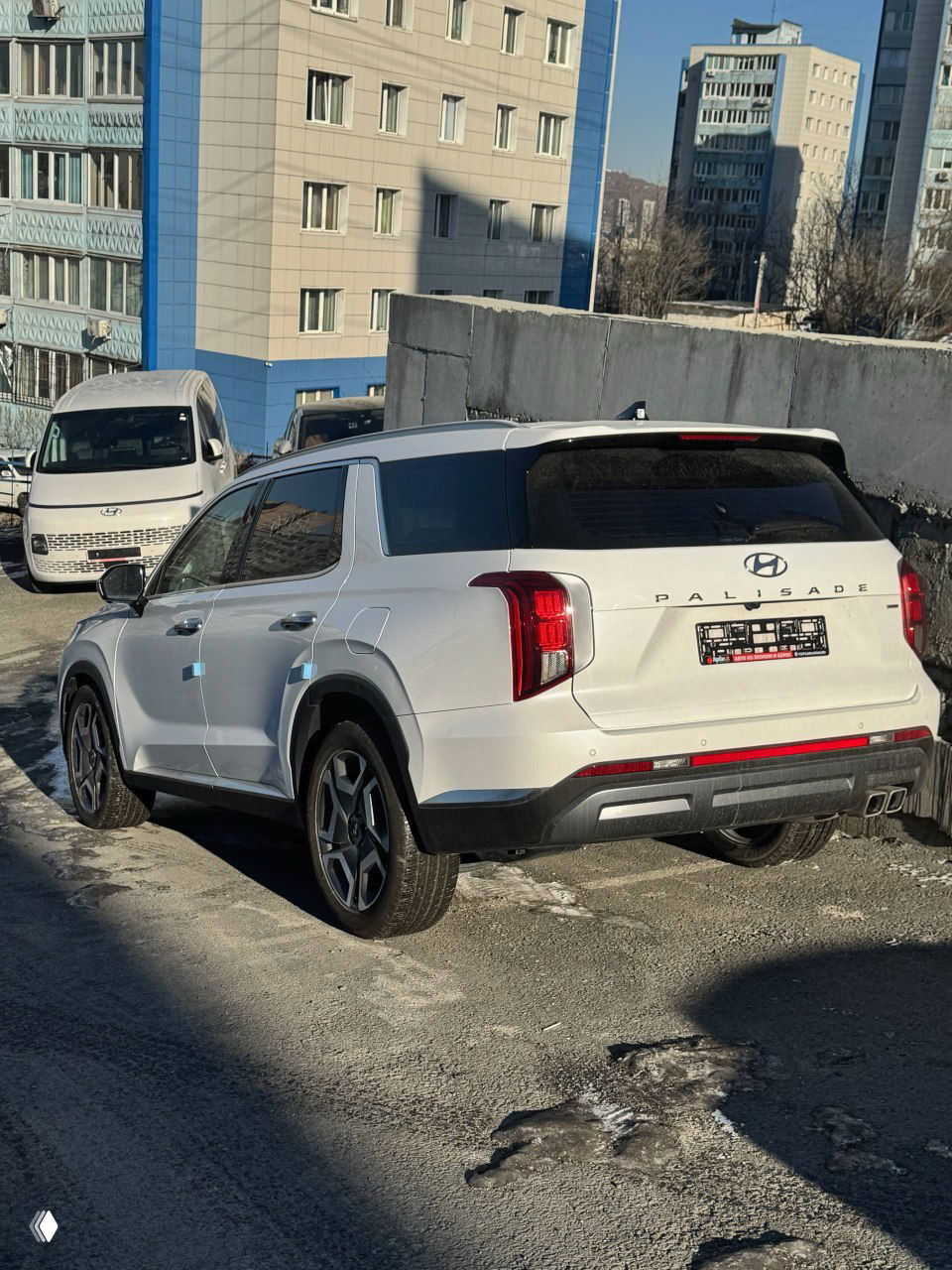 Вид сзади белого Hyundai Palisade рестайлинг, заметны эмблема PALISADE, задние фонари и номерной щиток, автомобиль на стоянке