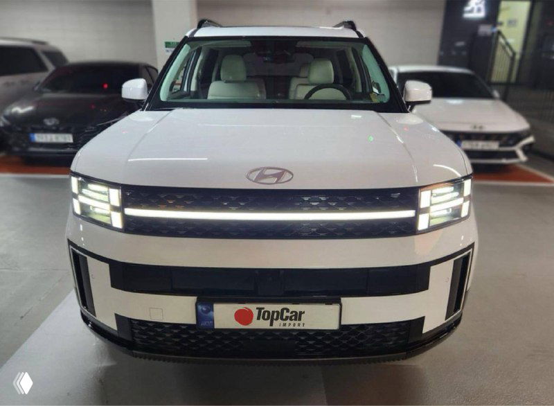 Фронтальный вид белого Hyundai Santa Fe (2024) в салоне-паркинге, крупный капот и современная светотехника спереди автомобиля.