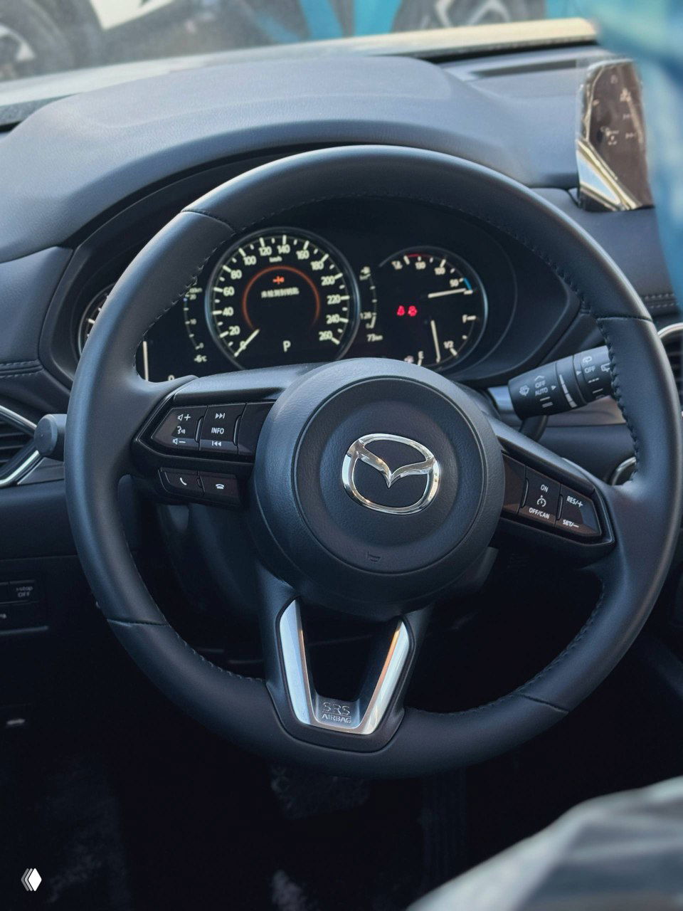 Крупный план рулевого колеса Mazda с логотипом, видна приборная панель и элементы управления мультимедиа в салоне автомобиля.