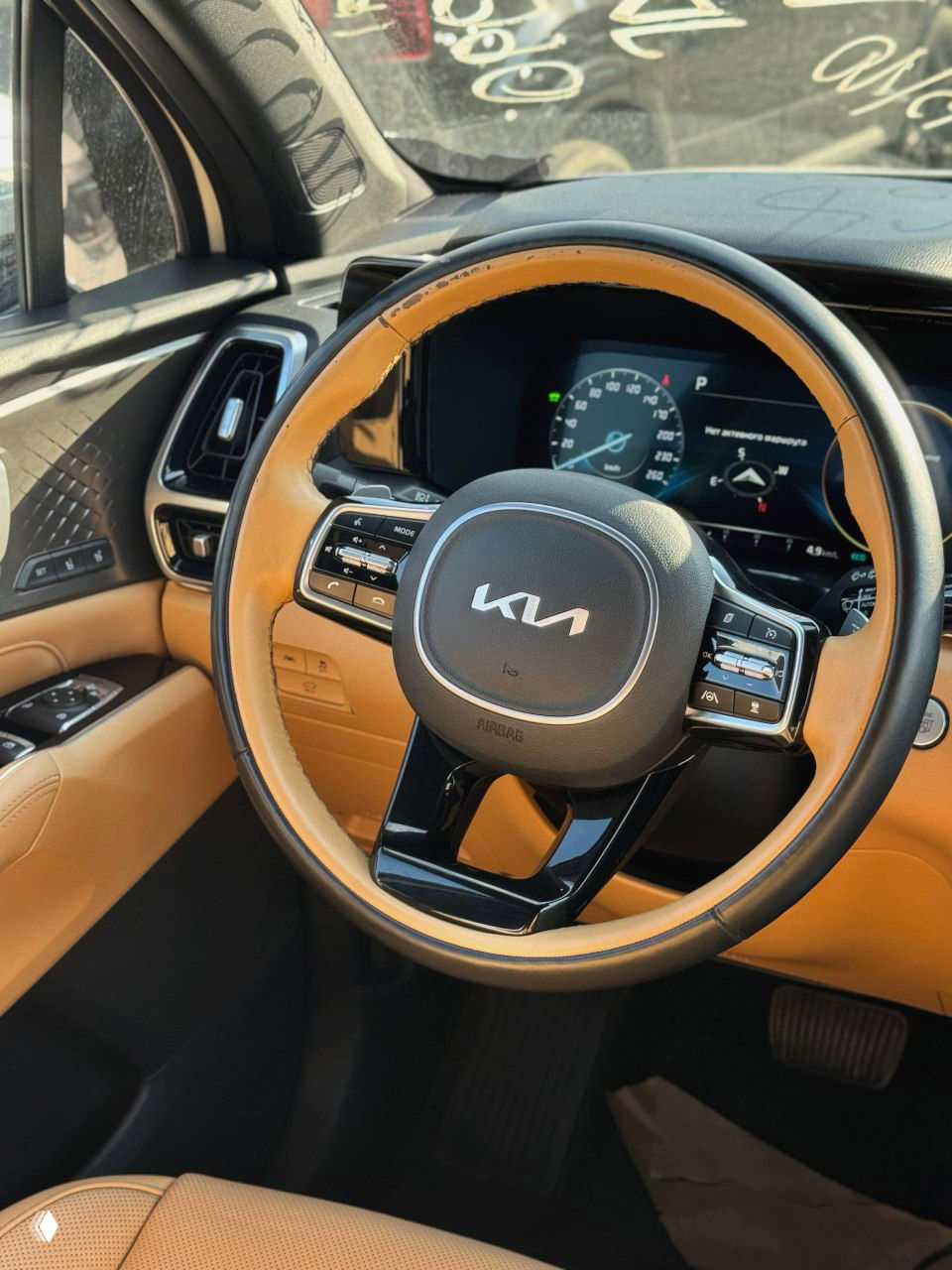 Крупный план рулевого колеса KIA с новым логотипом; двухцветная кожаная оплётка руля и вид на цифровую приборную панель и часть салона автомобиля.