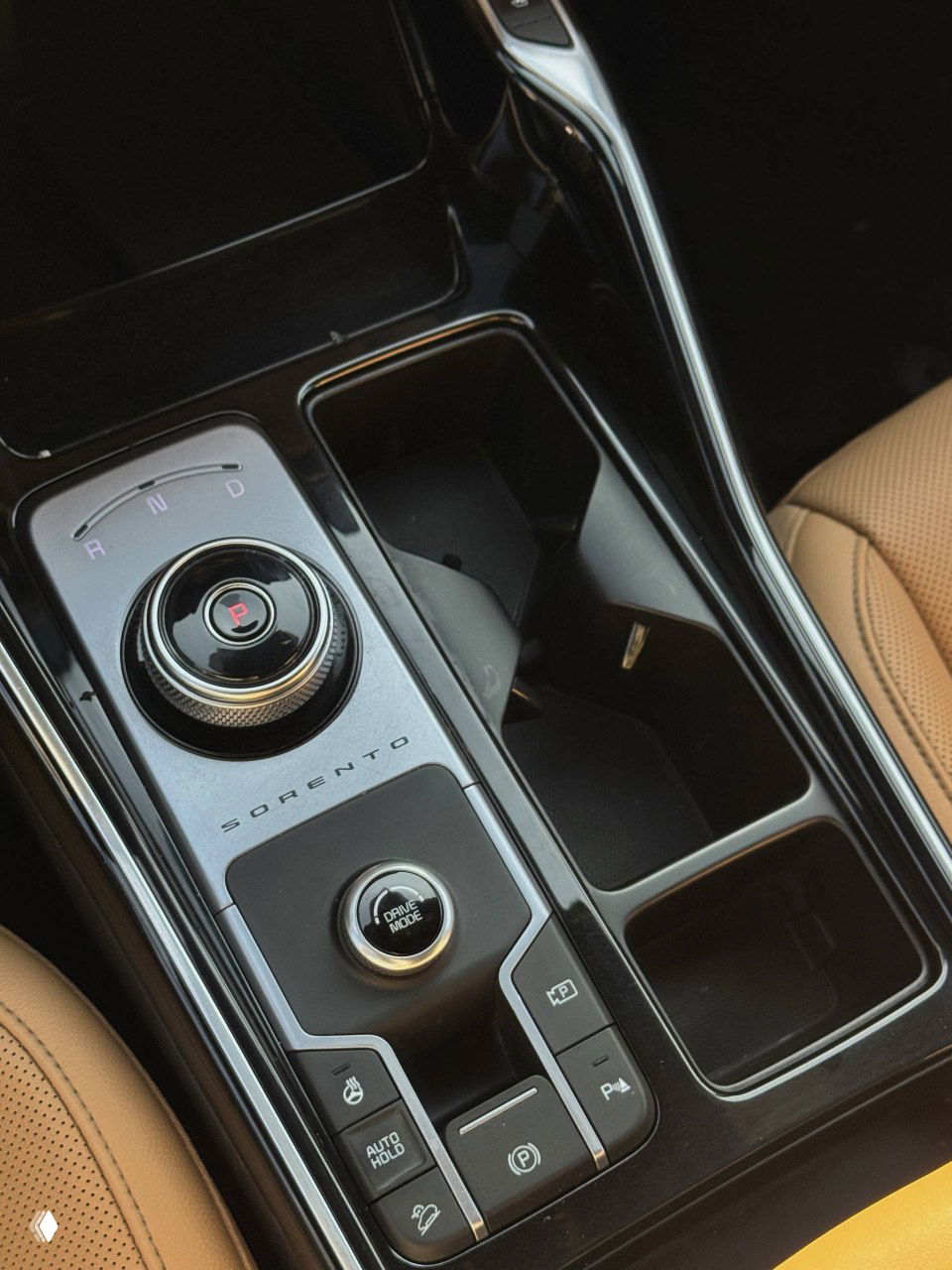 Ближайший план центральной консоли KIA Sorento: вращающийся селектор передач, обозначение SORENTO, кнопки режимов движения и держатели под кружки — аккуратный вид.