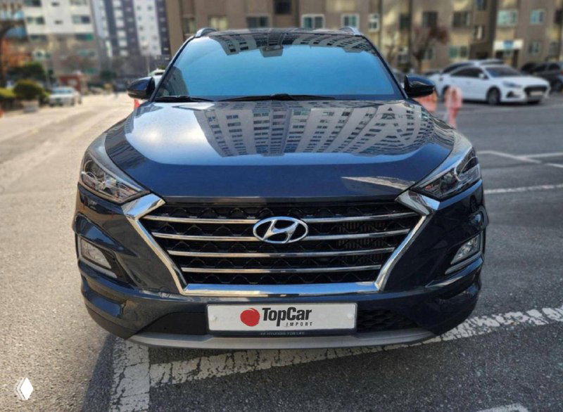 Hyundai Tucson тёмно-синего цвета, вид спереди на парковке; видно капот, решётку радиатора и отражение жилого дома в лаке.