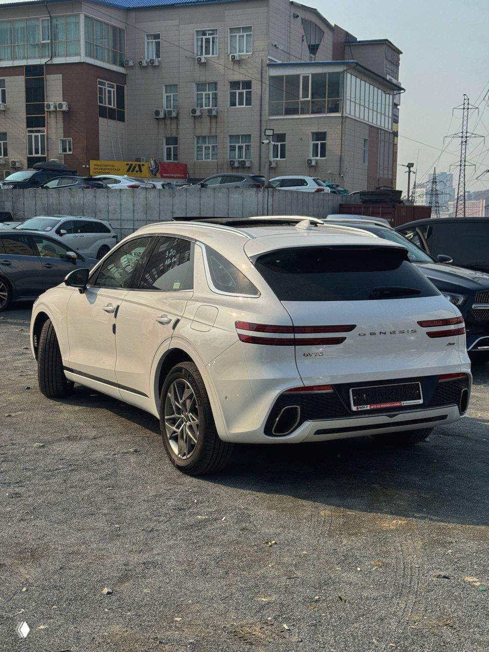 Genesis GV70 белого цвета, вид сзади и в три четверти на стоянке; заметны задняя оптика, надпись GENESIS и выхлопные насадки на фоне строений.