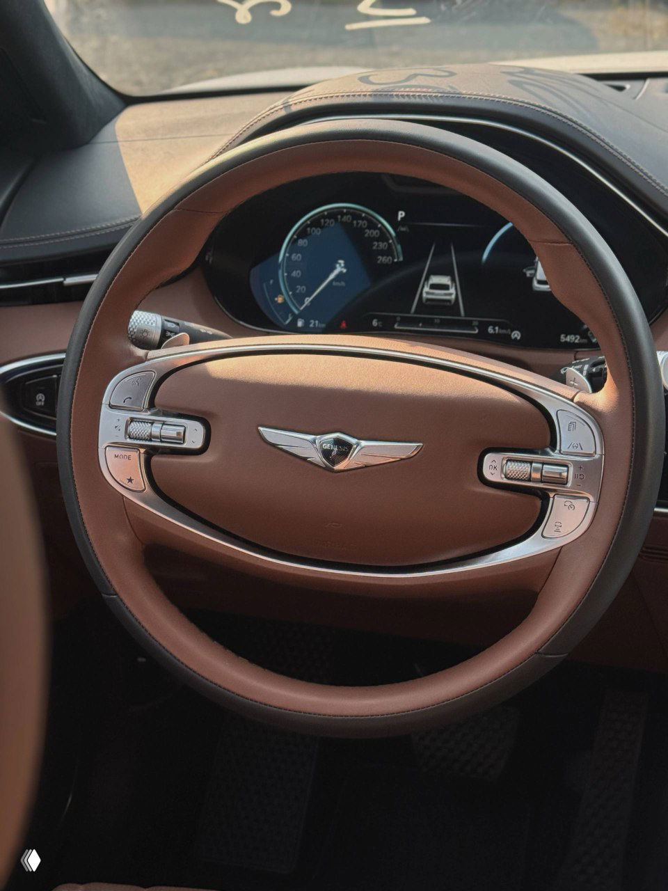 Крупный план руля Genesis GV70 — коричневая кожа, эмблема Genesis, элементы управления на стойках и цифровая панель приборов видны крупно.