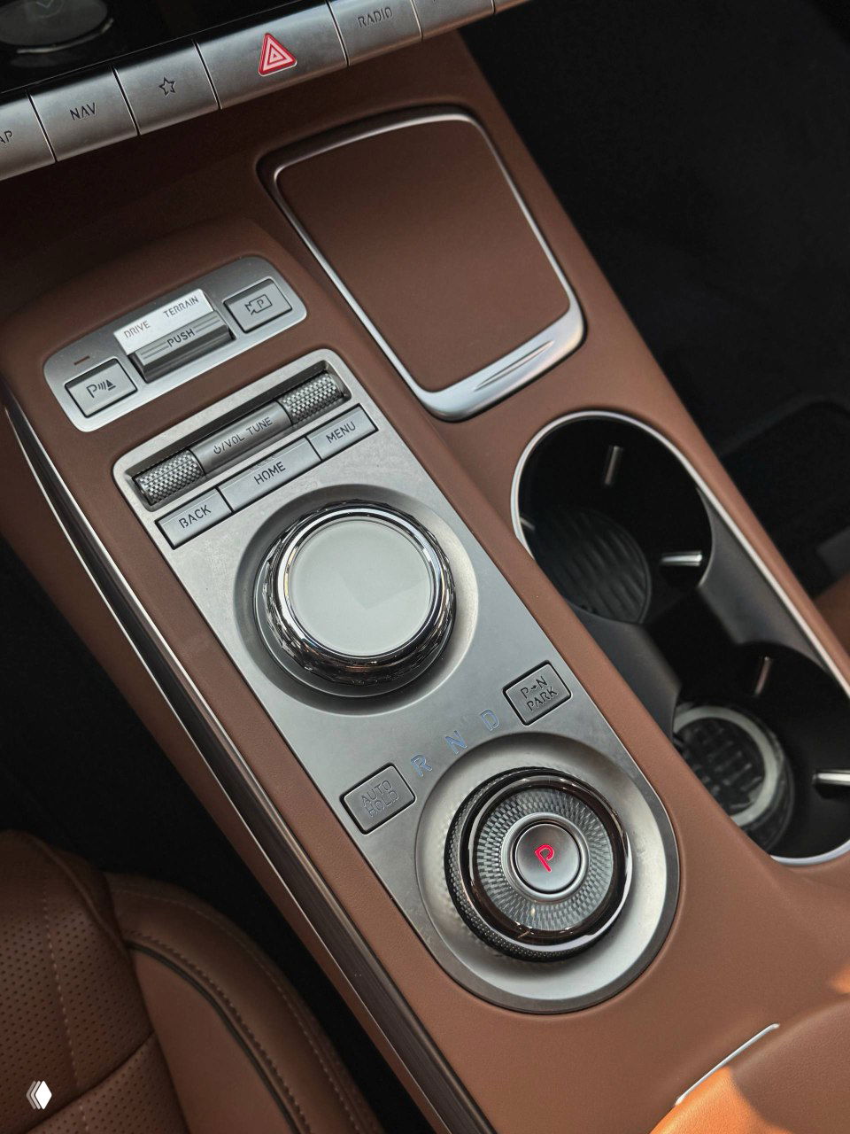 Крупный план центральной консоли Genesis GV70 с роликовым контроллером, металлическими вставками, подстаканниками и кожаным окантованием в сочетании коричневой кожи.