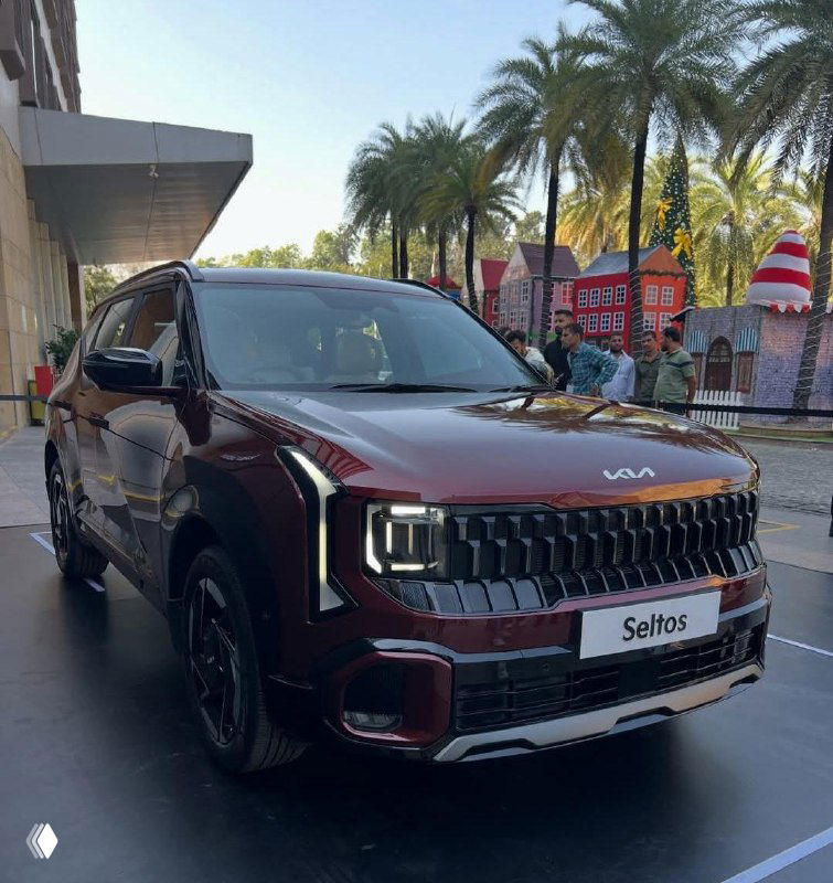 KIA Seltos 2026 бордового цвета выставлен в зале, фронтальный угол и фирменная решётка на фоне пальм и городской экспозиции.