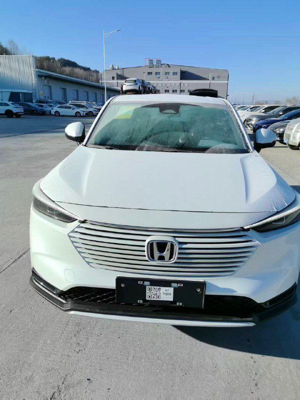 Фронтальный вид белого Honda Vezel 2026 с закрытой регистрацией, видны фары и капот, машина припаркована на складе экспорта.