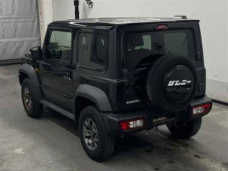 Задний ракурс чёрного Suzuki Jimny Sierra в складском боксе: запасное колесо на двери, задние фонари и номер с японской регистрацией на фото.