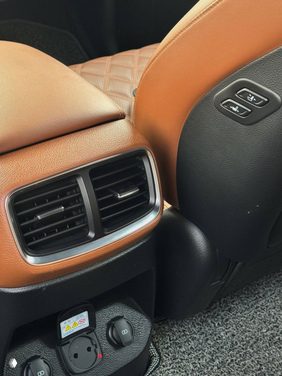 Крупный план заднего вентиляционного дефлектора и розетки питания в салоне KIA Mohave; показаны розетка и элементы комфорта для пассажиров.