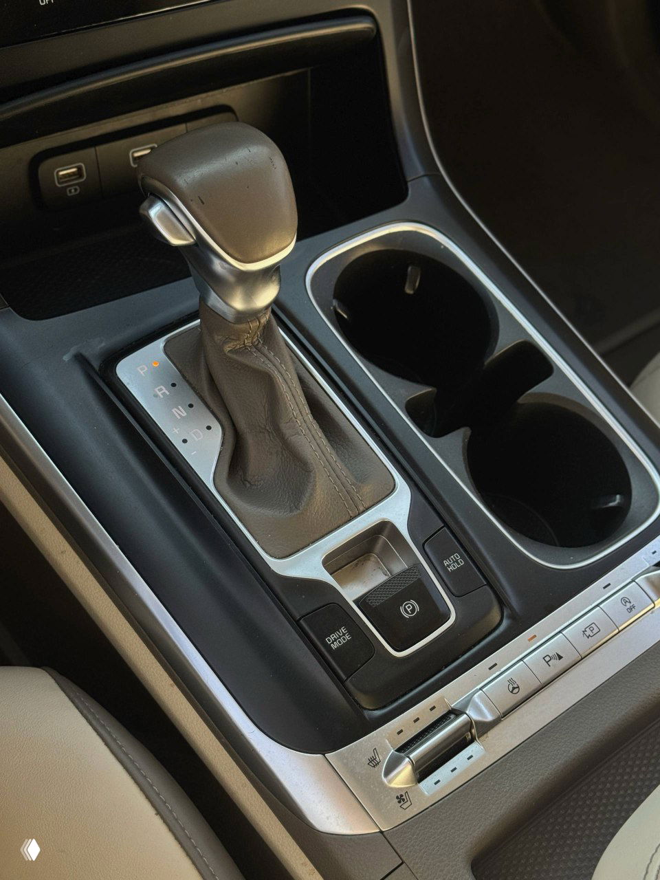 Селектор АКПП и центральная консоль KIA Carnival: ручка переключения, подстаканники и кнопки управления обогревом и режимами движения.