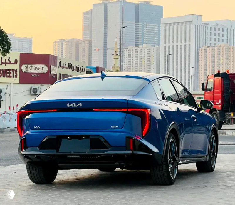 KIA K4 — задняя часть автомобиля: светодиодные фонари, фирменный логотип KIA и задний бампер на фоне городской застройки и грузового транспорта.