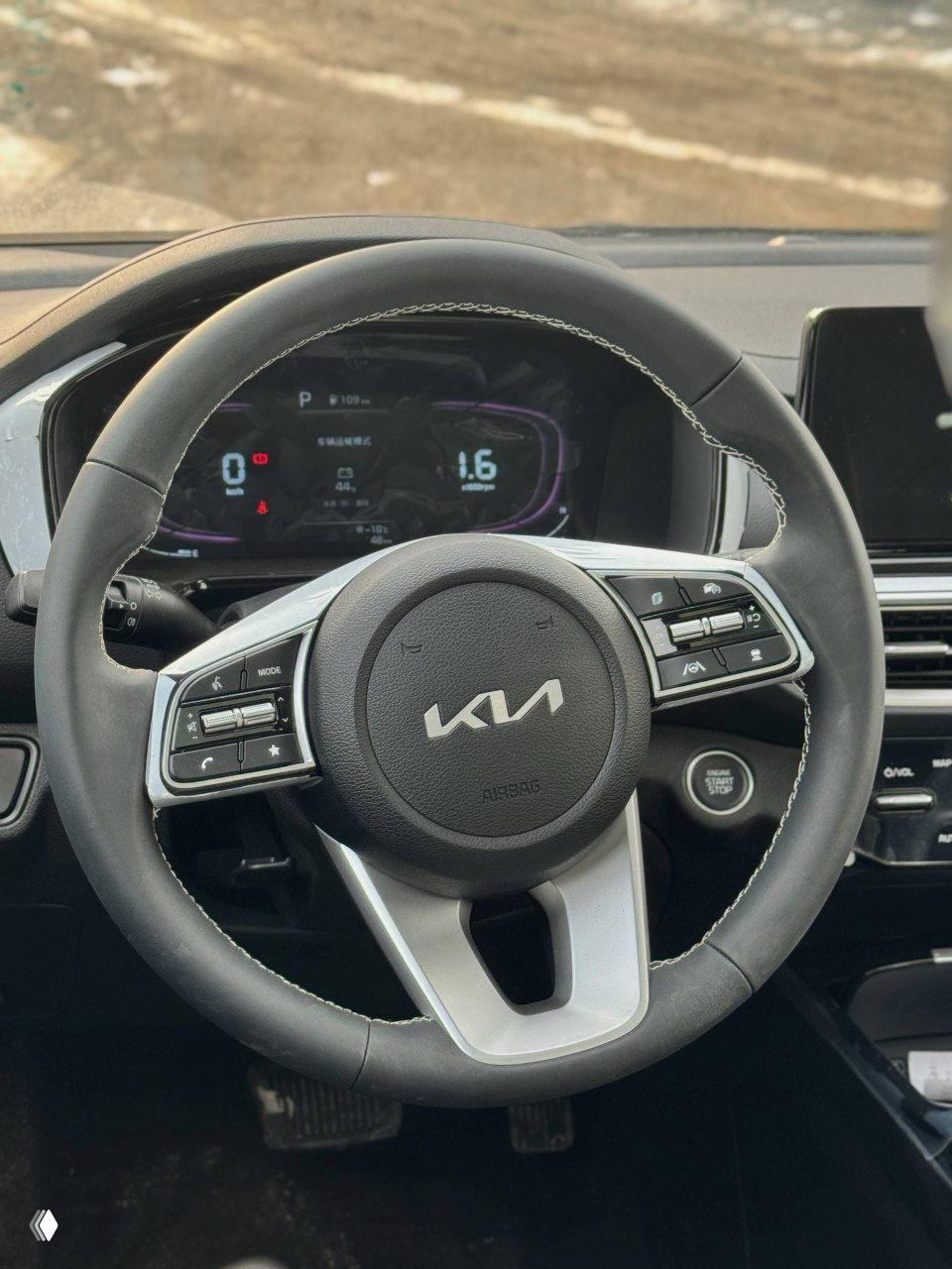 Крупный план рулевого колеса KIA с новой эмблемой, элементы управления на руле и цифровая приборная панель на фоне салона.