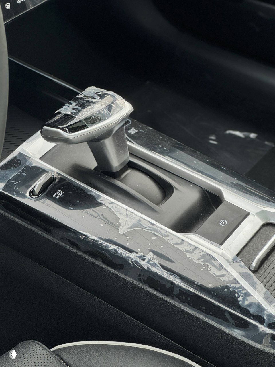 Крупный план селектора автоматической трансмиссии KIA K3 с защитной плёнкой на элементах отделки, вид на подлокотник и передний ряд кресел.