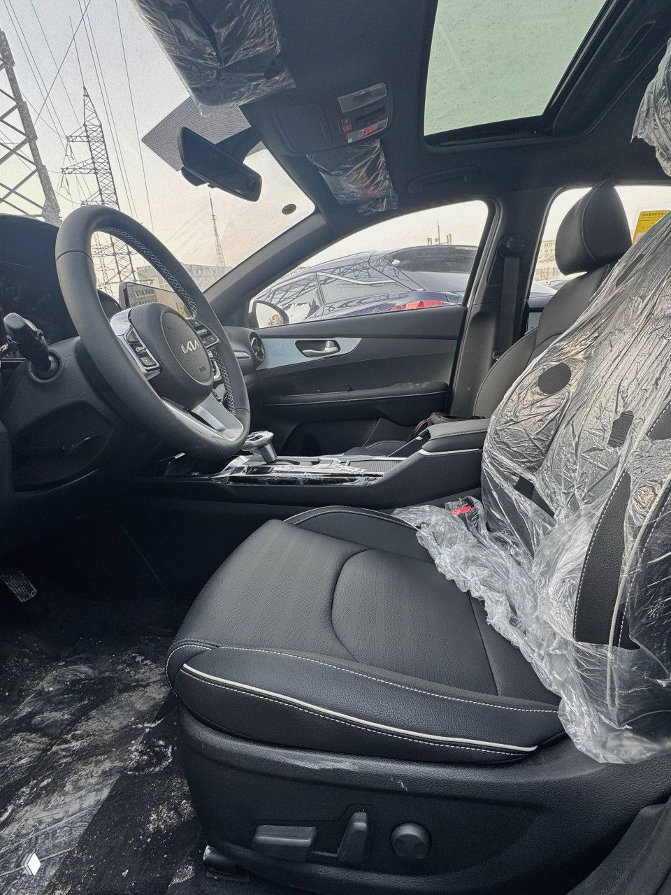 Вид на переднее водительское кресло и панорамный потолок KIA K3, кресло в защитной плёнке и интерьер в чёрной обивке.