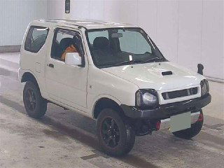 Белый Suzuki Jimny 2011, вид спереди справа в ангаре аукциона; тёмные диски, заметен небольшой тюнинг и общие следы эксплуатации.