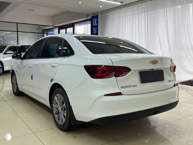 Задняя часть белого Chevrolet Cruze 2021 в салоне; вид на багажник, фон — занавеси и стенды автосалона, номер закрыт для экспозиции.