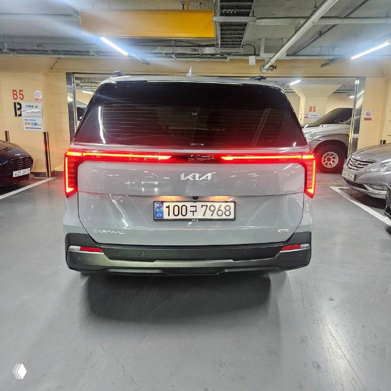 Задняя часть белого KIA Carnival 2025 с характерной светодиодной полосой и открытым багажником, парковка на фоне колонн.