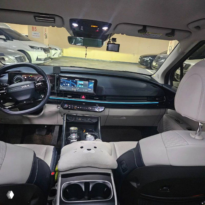 Панорамный вид торпедо и передних сидений KIA Carnival: большой сенсорный экран, мультируль и светлый салон с тканевой отделкой.