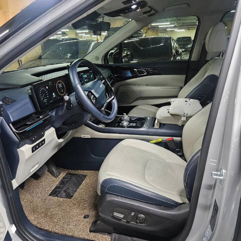 Вид водительского места KIA Carnival: мультируль, приборная панель и сиденье водителя с электроприводом в светлой обивке.