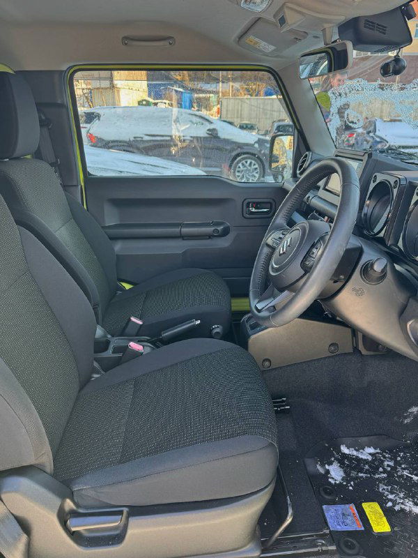 Вид на передние сиденья и пассажирскую часть салона Suzuki Jimny: тканевая обивка, сиденья, дверная панель и часть лобового стекла с наружным окружением на фоне.