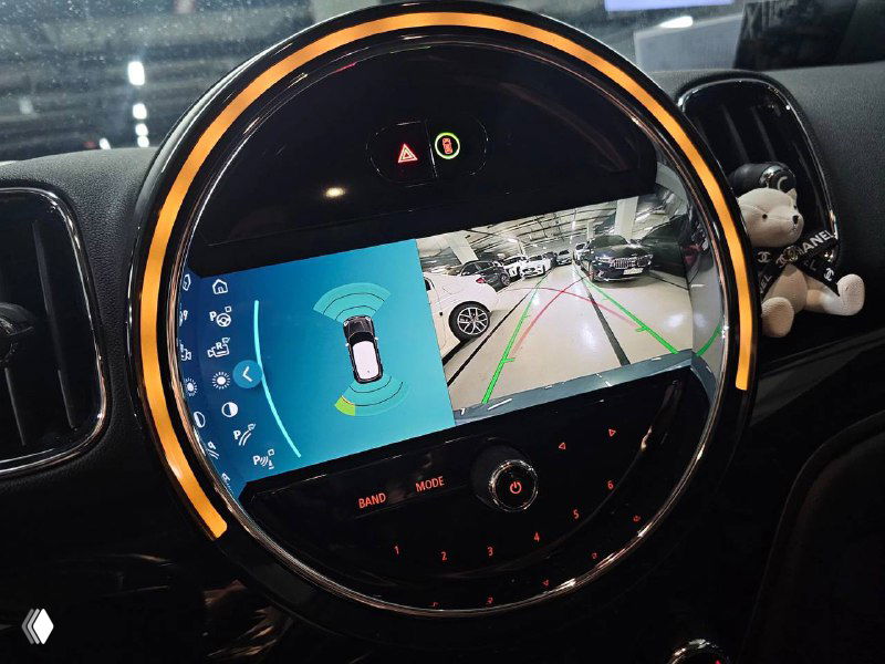 Крупный план центрального круглого дисплея Mini Cooper Countryman с активной камерой парковки и графикой помощи при маневрировании в паркинге.