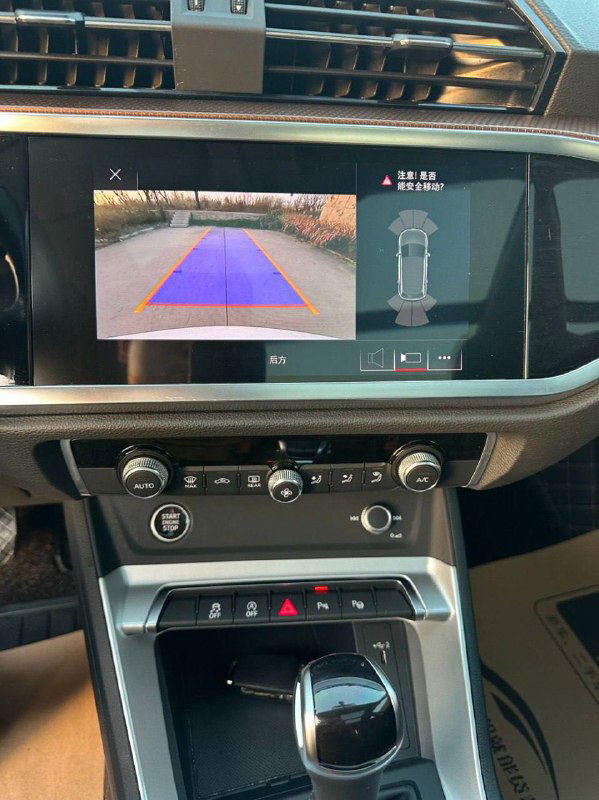 Экран мультимедиа Audi Q3 с включённой камерой заднего вида и разметкой парковки, панель климат‑контроля и элементы консоли, демонстрация техники и опций.