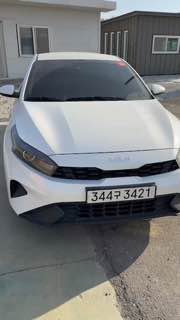 Фотография белого KIA K3 спереди на стоянке в Южной Корее; видны фары, решётка радиатора и корейский номер на асфальтовой поверхности рядом с бытовками.