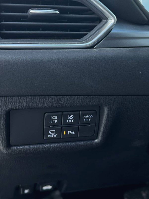 Фрагмент панели с кнопками управления TCS, обзора и парковки Mazda CX-5 2025, пластиковая отделка и кнопки системы помощи водителю видно крупным планом.